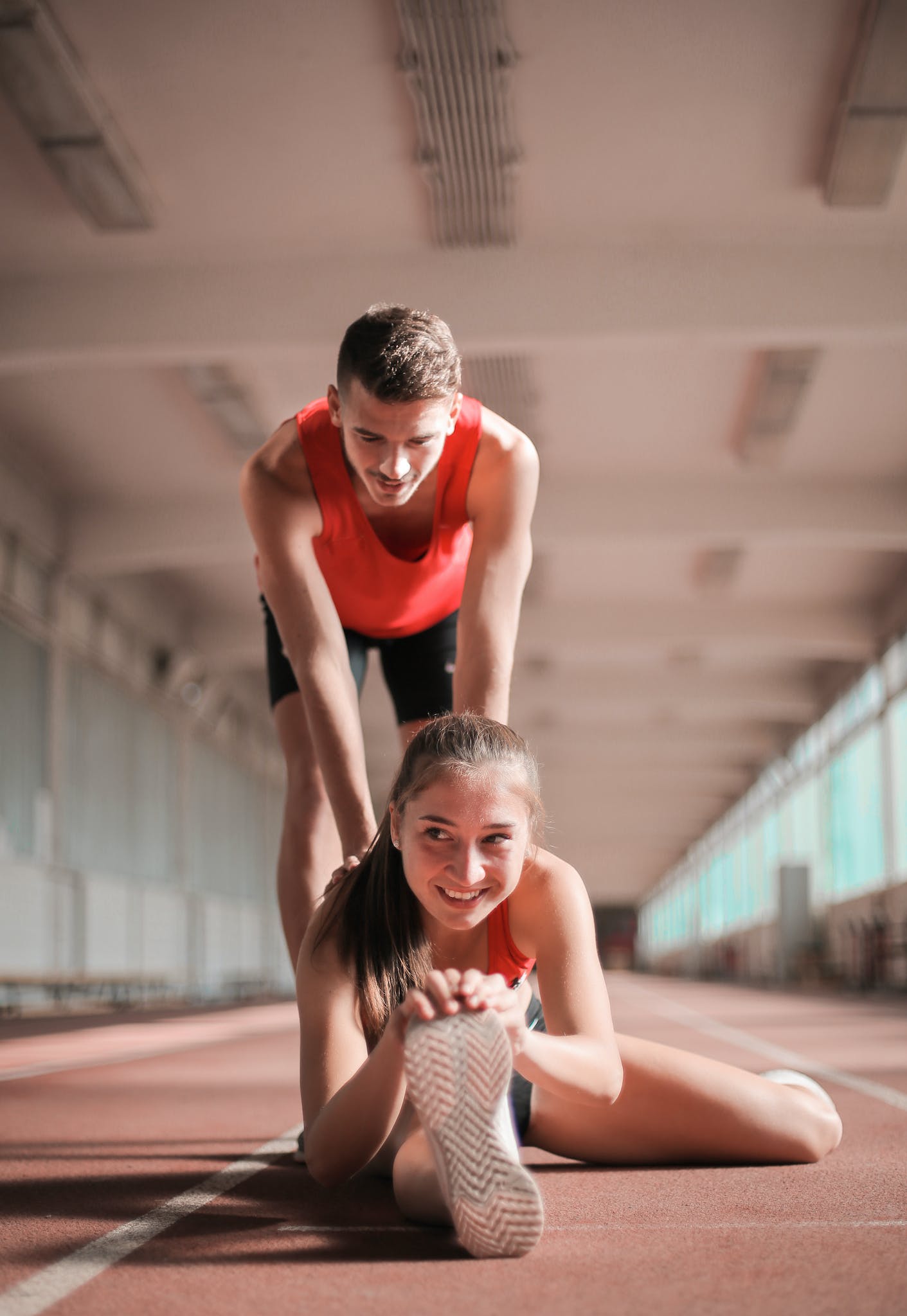 Cheerful sportswoman sitting on floor and stretching legs while man pushing back forward helping with warm up